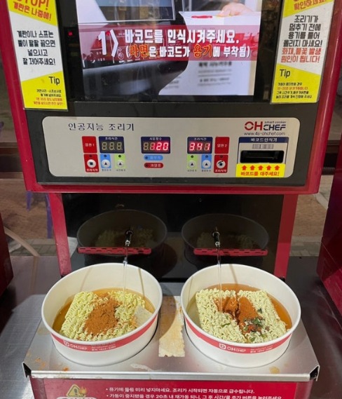 An automatic ramyeon cooking machine at a Han River convenience store with two paper bowls of noodles ready to cook