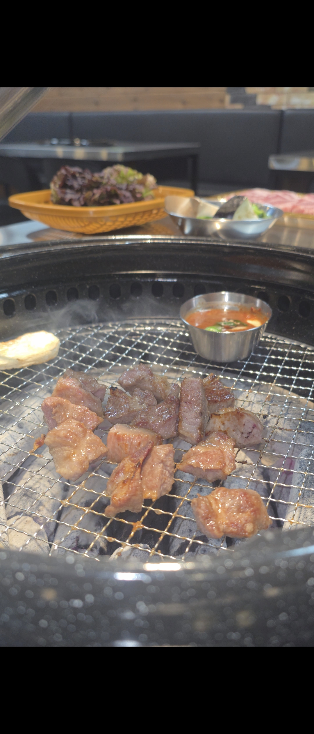 Bite-size meat grilling over charcoal on a wire-mesh grill, with a cup of dipping sauce