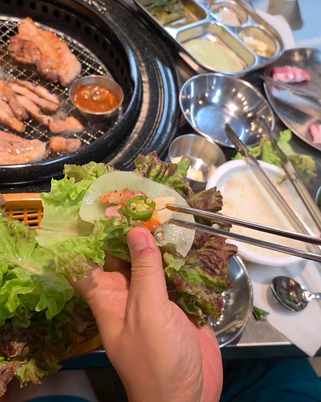 A hand holding a small samgyeopsal ssam — grilled pork belly, sauce, and chili wrapped in a lettuce leaf at a Korean BBQ table