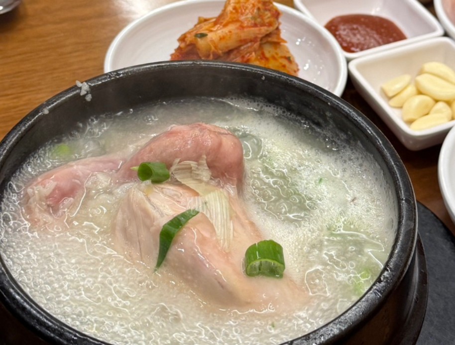 A steaming stone bowl of samgyetang on a restaurant table, with kimchi, gochujang and a dish of whole garlic cloves set behind it