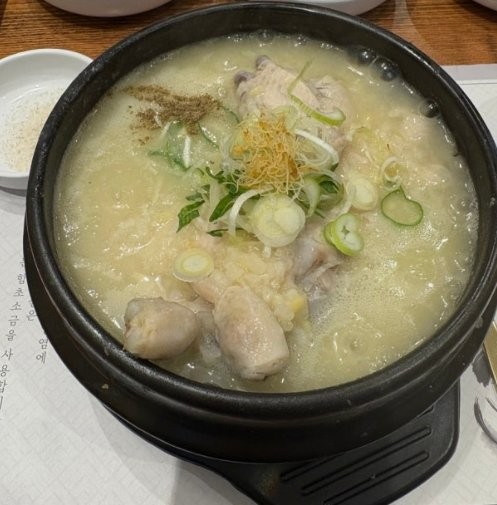 A bowl of samgyetang topped with sliced spring onion and shredded ginseng, with a small dish of salt and pepper beside it for seasoning the broth