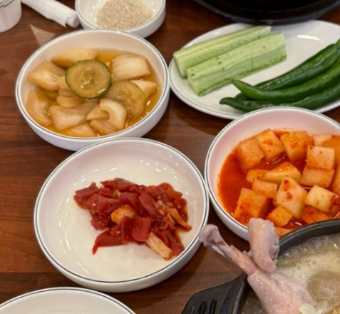 The side dishes served alongside samgyetang — pickled radish, cucumber sticks and green chillies, gochujang, and cubed radish kimchi (kkakdugi) — surrounding the edge of the soup bowl